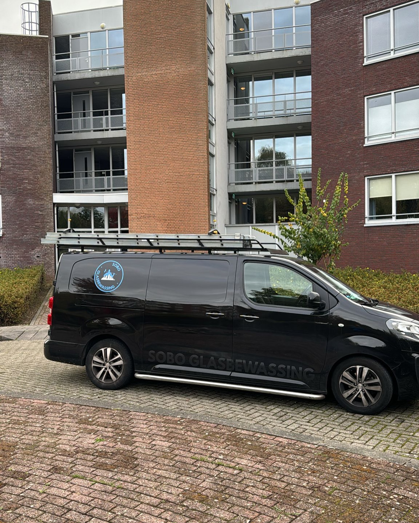 Zwarte bedrijfsbus van Sobo glasbewassing met ladders op het dak, voor een appartementencomplex